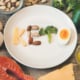 tavola con vari ingredienti che circondano un piatto in cui c'è scritto keto con formaggio, frutta secca broccolo e uovo