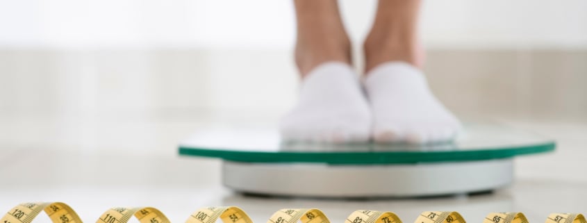 in primo piano c'è un metro da sarta. Sullo sfondo i piedi di una persona con i calzini, su una bilancia